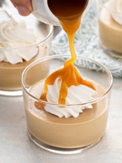 Caramel sauce being poured over a dish of butterscotch pudding topped with whipped cream.