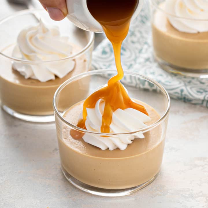 Caramel sauce being poured over a dish of butterscotch pudding topped with whipped cream.