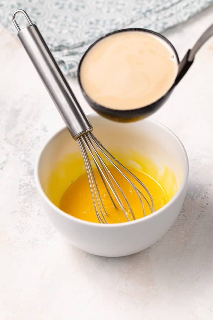 Hot milk and sugar about to be poured into whisked egg yolks in a small white bowl.