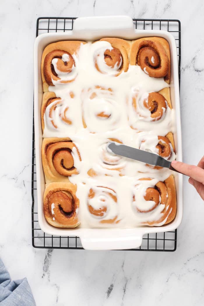 Spatula spreading icing on top of overnight cinnamon rolls in a white baking dish