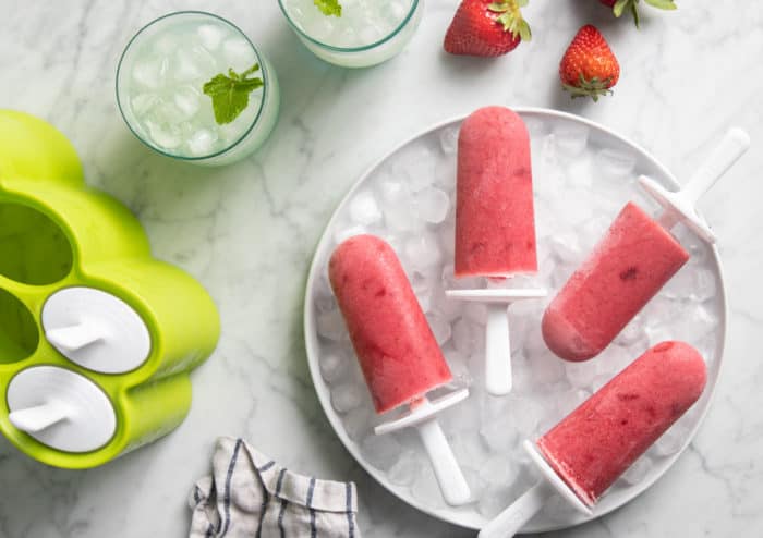White bowl of ice with strawberry popsicles on top set next to a popsicle mold.