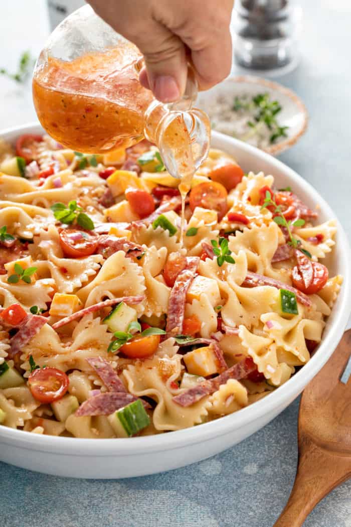 Dressing being drizzled over italian pasta salad in a white serving bowl.