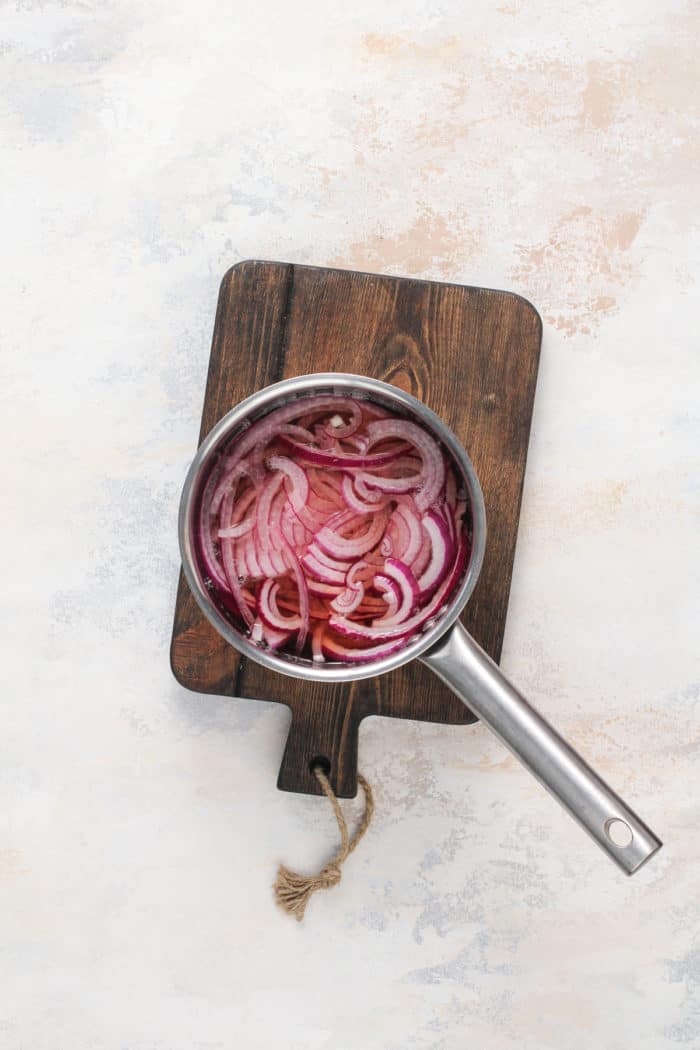 Red onions marinating in vinegar brine in a saucepan set on a wooden board.