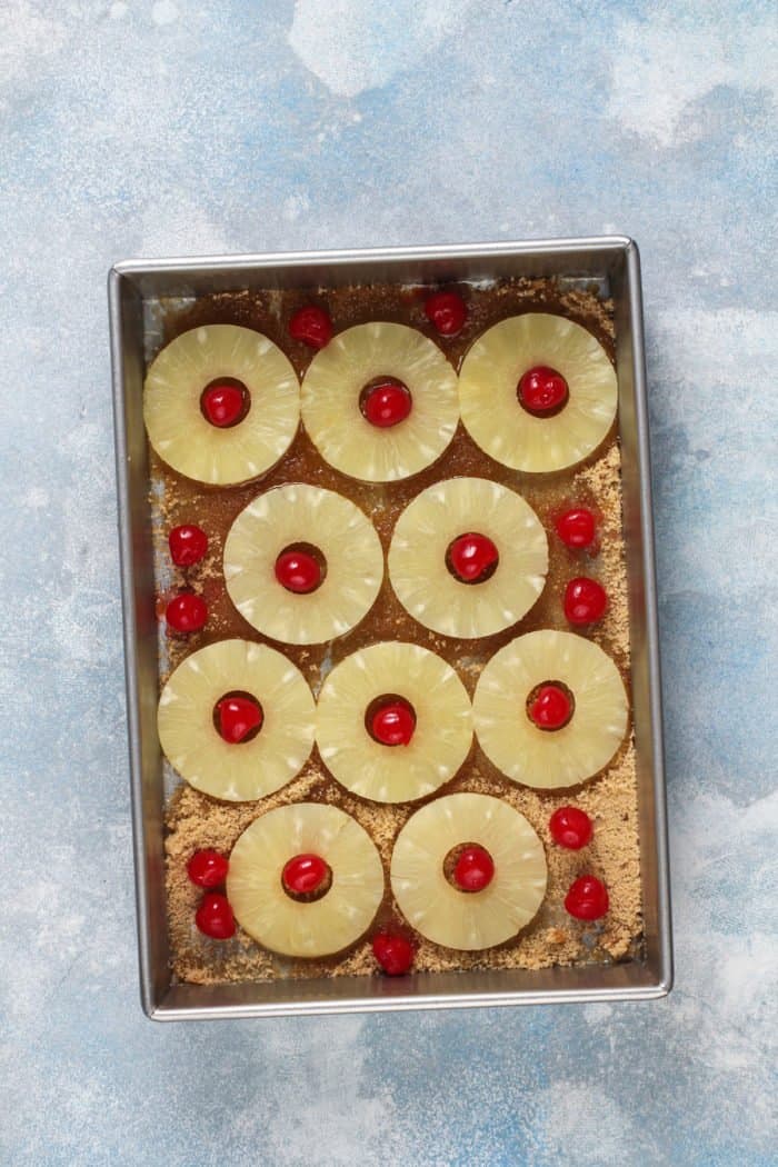 Pineapple slices and maraschino cherries arranged in the bottom of a cake pan.