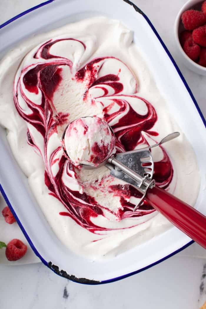 Ice cream scoop serving up a scoop of swirled berry no-churn ice cream from a freezer container.