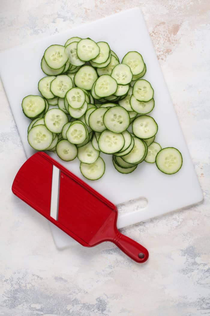 Sliced cucumbers next to a mandoline slicer on a white cutting board.