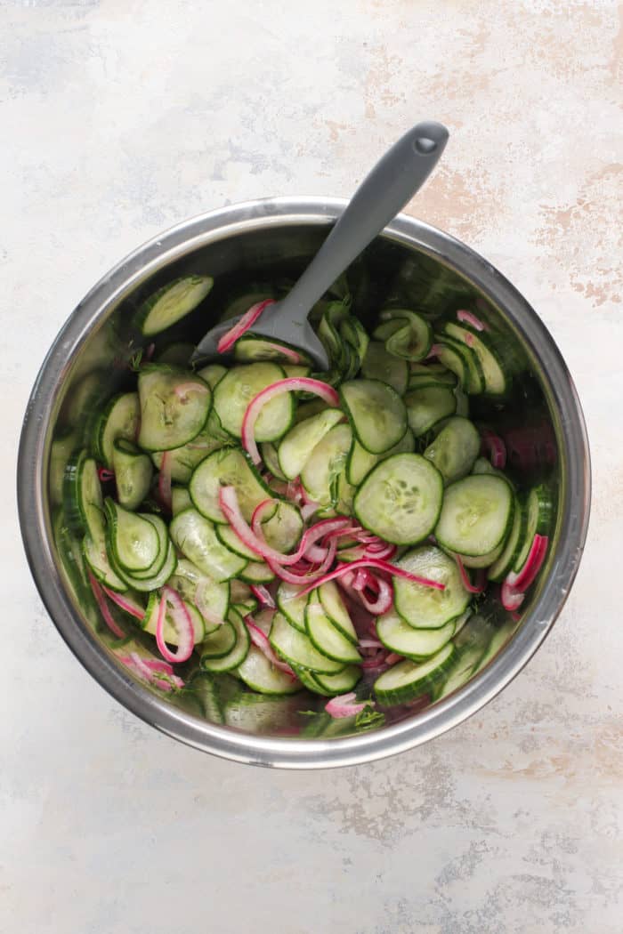 Cucumber salad being stirred with a spatula in a metal mixing bowl.
