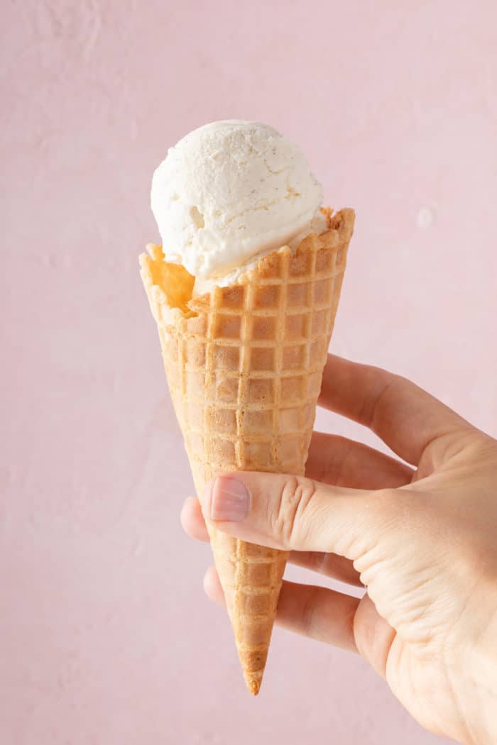 Hand holding up a waffle cone with a scoop of vanilla no-churn ice cream in front of a pink wall.