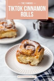 Frosted cinnamon roll with heavy cream on a white plate. A second cinnamon roll and a cup of coffee are in the background. Text overlay includes recipe name.