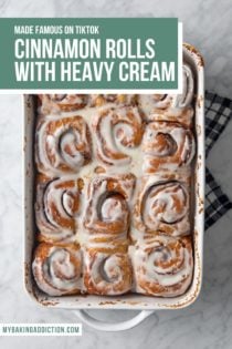 Frosted cinnamon rolls with heavy cream in a white baking dish, resting on a tea towel to cool. Text overlay includes recipe name.