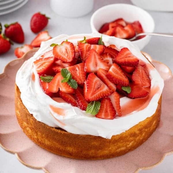 Pink cake plate holding a strawberry shortcake topped with whipped cream and sliced strawberries.