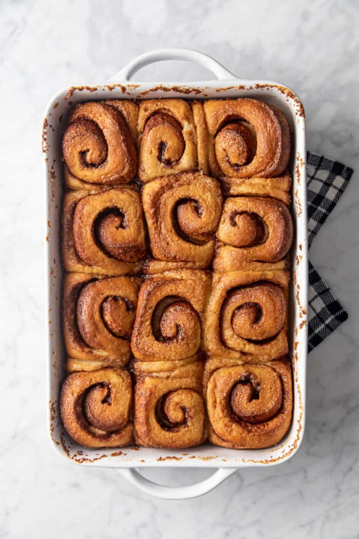 Baked cinnamon rolls with heavy cream in a white baking dish, set on a tea towel to cool.