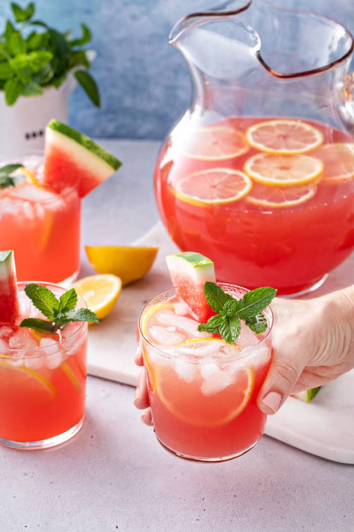 Hand holding up a glass of watermelon lemonade. More glasses and a pitcher of the lemonade are in the background.