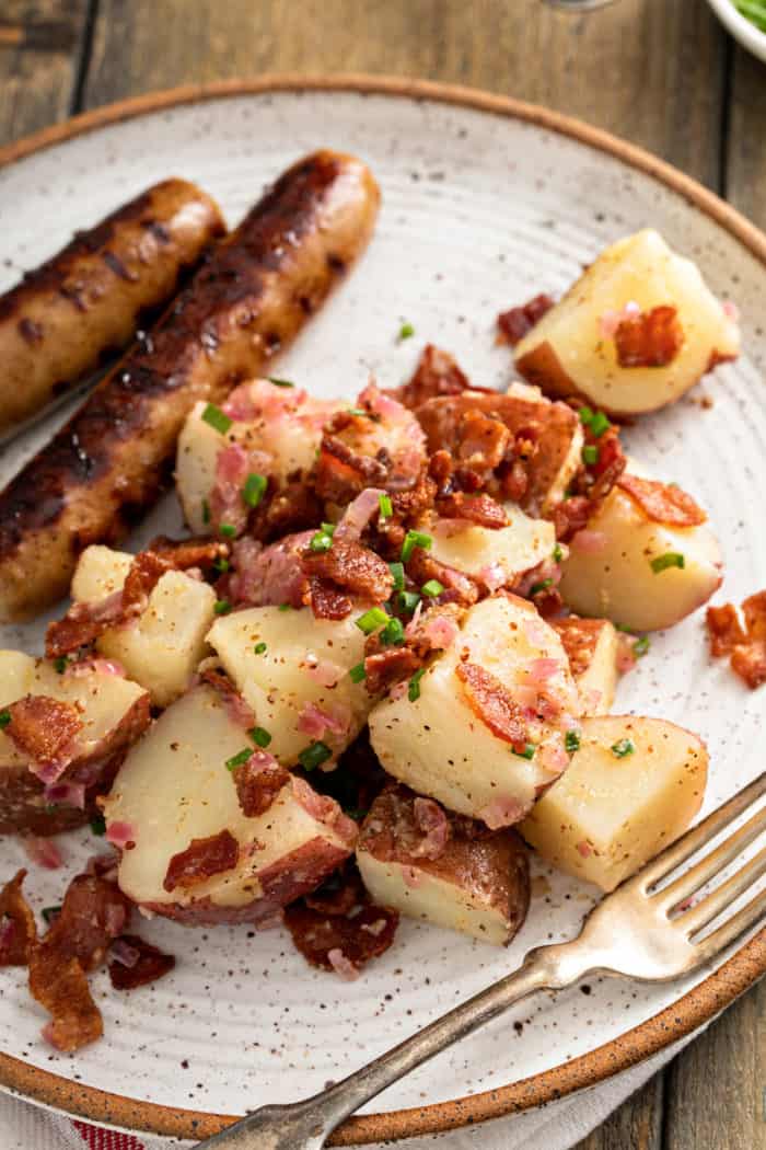 Close up view of german potato salad on a plate. Two brats are just visible off to the side of the salad.