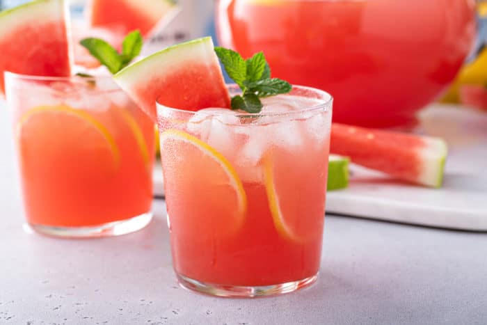 Glass of watermelon lemonade on a gray countertop. There are more glasses and a pitcher of watermelon lemonade in the background.