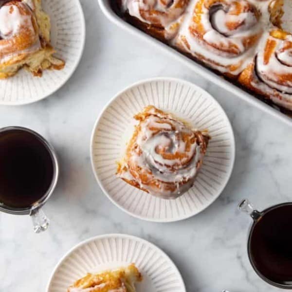 Three white plates with tiktok cinnamon rolls set on a marble countertop next to a baking dish of cinnamon rolls.