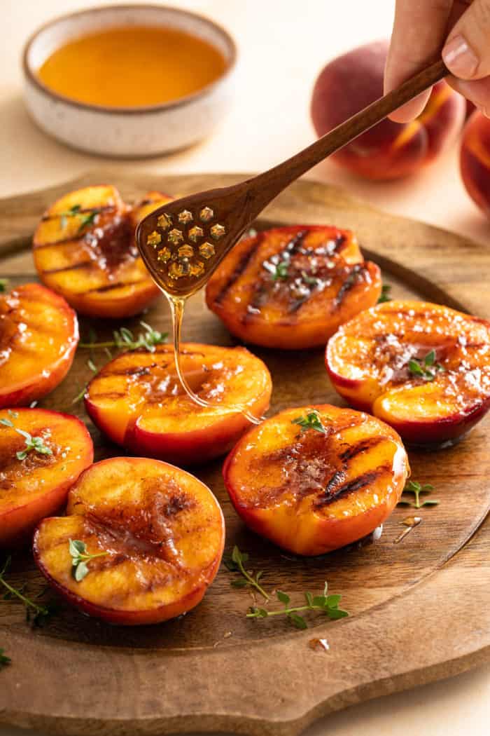 Honey being drizzled over grilled peach halves arranged on a wooden platter.