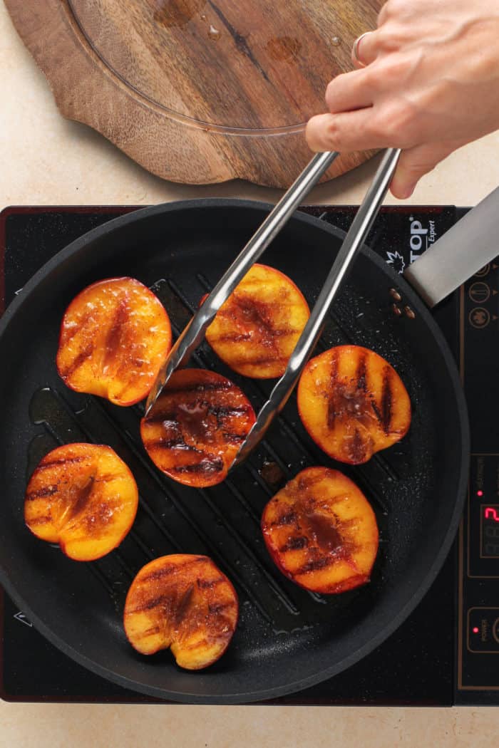 Tongs flipping over grilled peach halves in a grill pan.