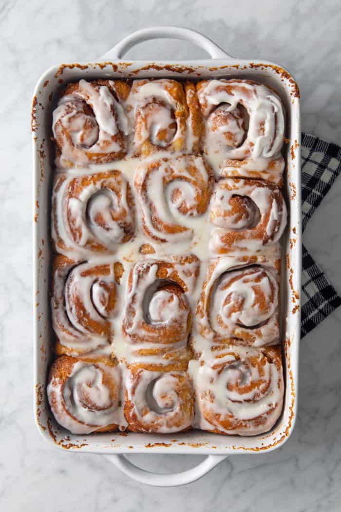 Frosted cinnamon rolls with heavy cream in a white baking dish, resting on a tea towel to cool.