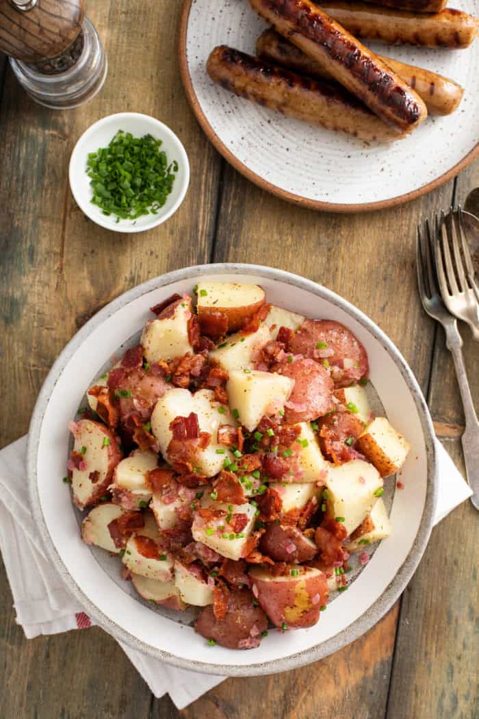 Overhead view of a serving bowl of german potato salad set on a wooden table next to a plate of grilled brats.