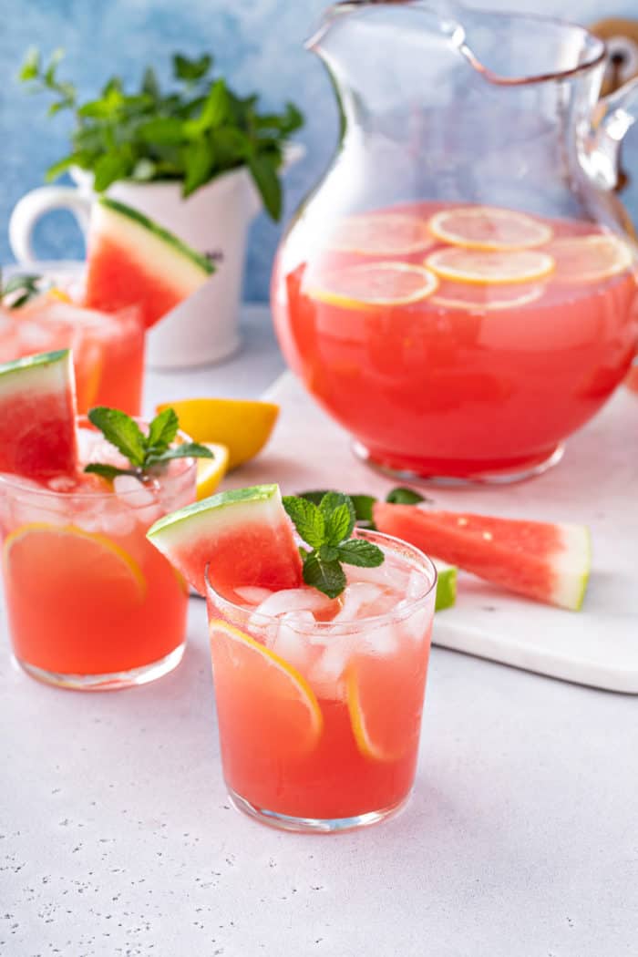Glasses of watermelon lemonade on a gray countertop with a pitcher of the lemonade in the background.