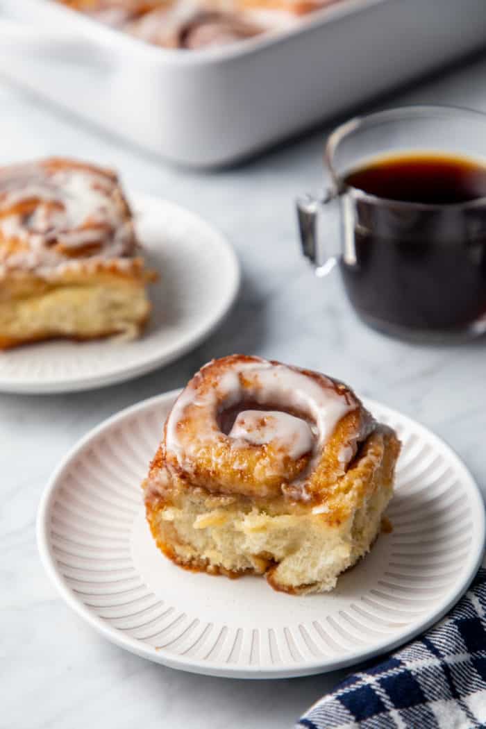 Frosted cinnamon roll with heavy cream on a white plate. A second cinnamon roll and a cup of coffee are in the background.