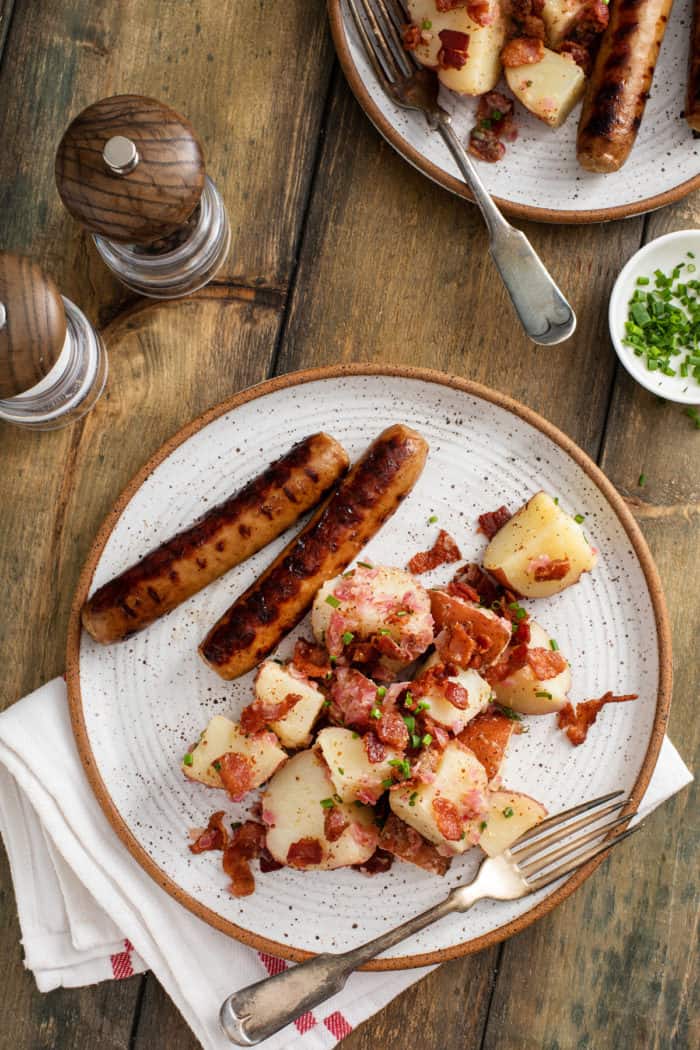 Overhead view of a pottery plate on a wooden table. Two brats and german potato salad are on the plate.