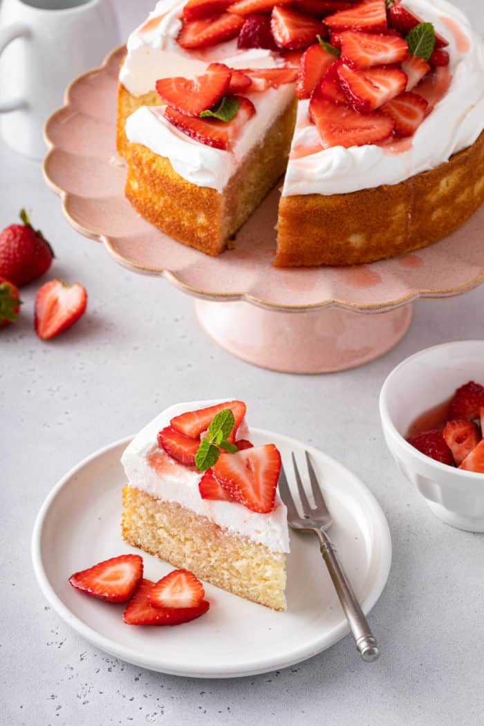 White plate with a slice of strawberry shortcake on it set next to a pink cake stand with the whole cake on it.