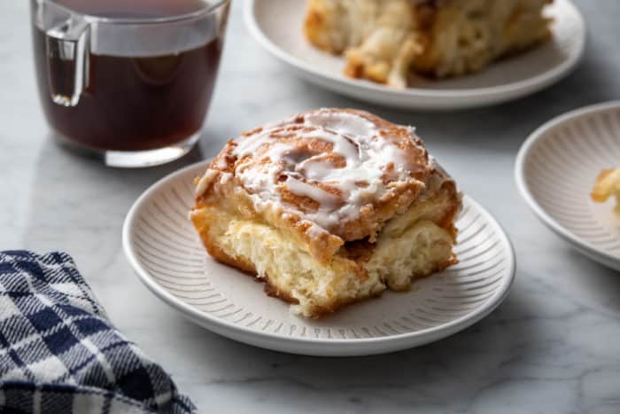 White plate holding a tiktok cinnamon roll. A cup of coffee and a second cinnamon roll are in the background.