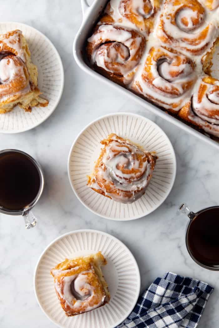 Three white plates with tiktok cinnamon rolls set on a marble countertop next to a baking dish of cinnamon rolls.