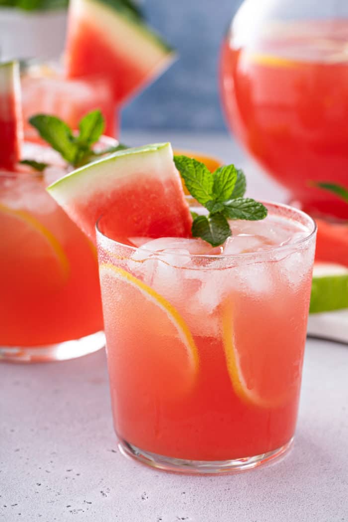Close up of watermelon lemonade in a glass, garnished with a slice of watermelon, lemon slices, and mint.