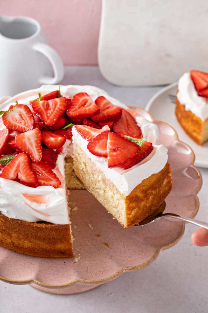 Cake server lifting up a slice of strawberry shortcake from the full cake on a pink cake plate.