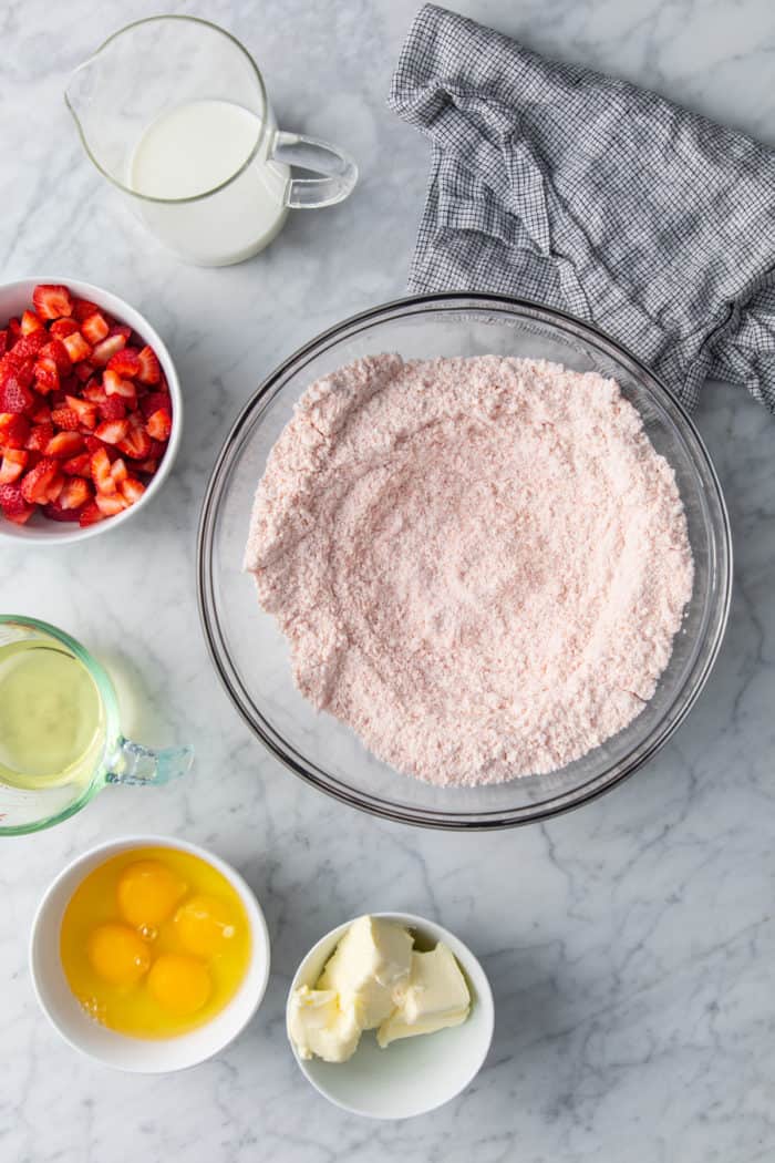 Cake mix, jello mix, and butter combined in a glass mixing bowl, with the other ingredients for strawberry cake surrounding it.