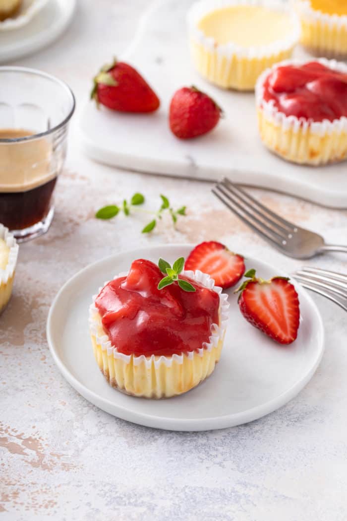 Mini cheesecake topped with strawberry pie filling on a white plate next to a halved strawberry.