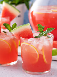 Glasses of watermelon lemonade on a counter in front of a pitcher of watermelon lemonade.