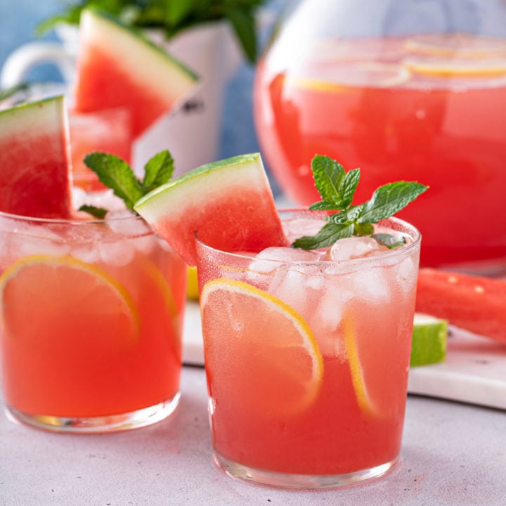 Glasses of watermelon lemonade on a counter in front of a pitcher of watermelon lemonade.