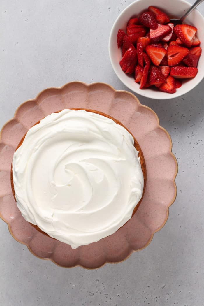 Yellow sponge cake topped with whipped cream on a pink cake plate next to a bowl of sliced strawberries.