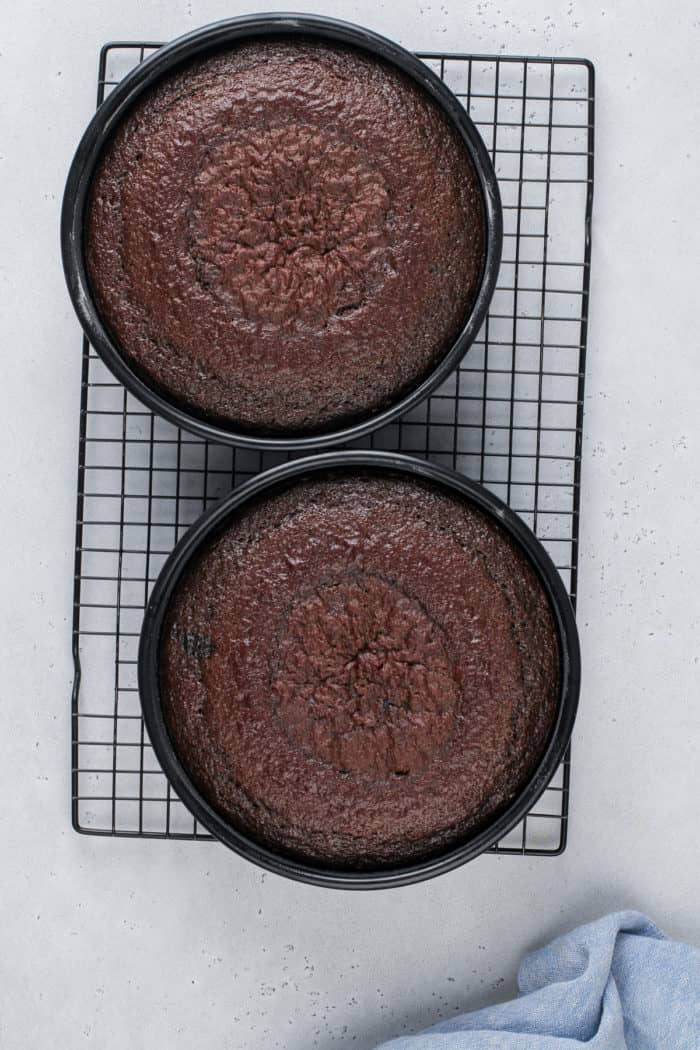 Baked black magic cake in two round cake pans, both set on a wire cooling rack.