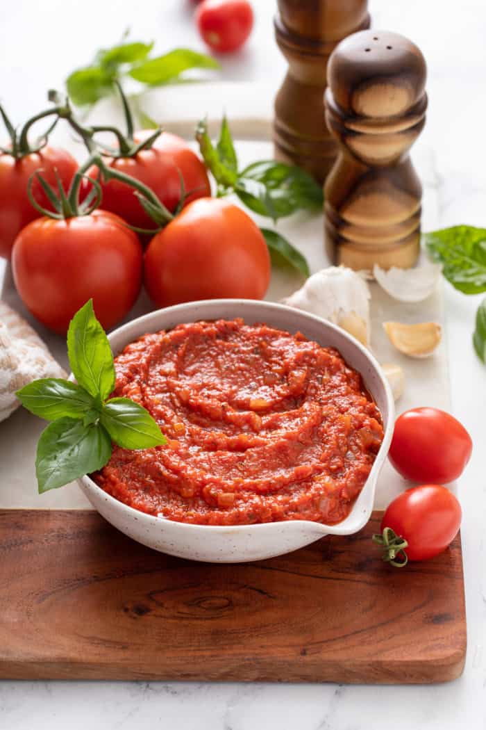 White bowl filled with marinara sauce, set on a wooden and marble board and surrounded by tomatoes and basil.