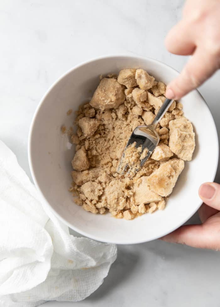 Hand using a fork to break up clumps of brown sugar in a white bowl.