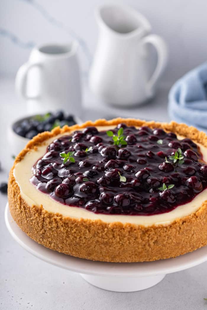 Close up of a cheesecake topped with blueberry sauce, set on a white cake plate.