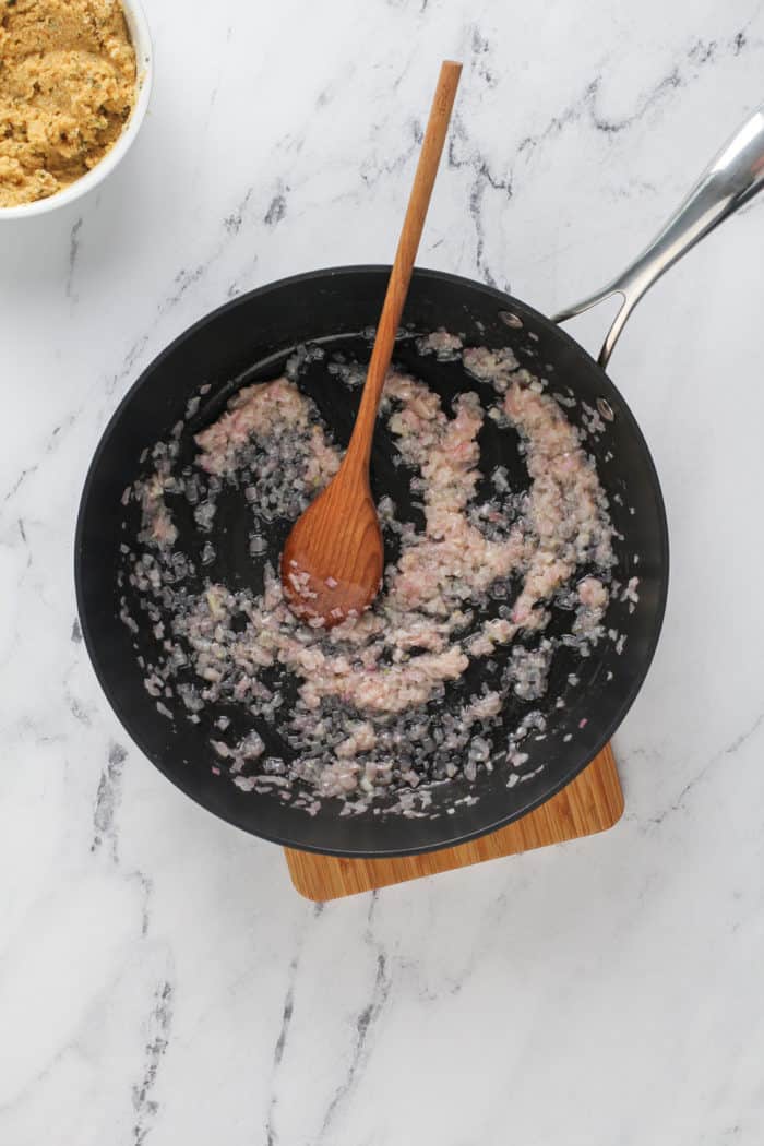 Cooked shallots and garlic being stirred in a black skillet with a wooden spoon.