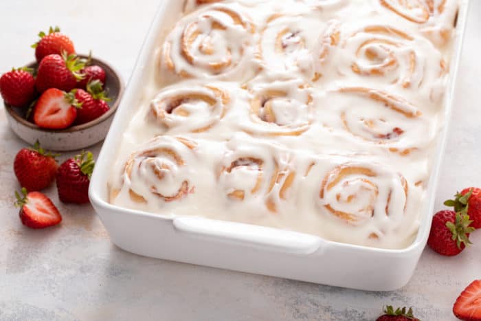 Frosted strawberry rolls in a white baking dish, set on a marble countertop next to fresh strawberries.