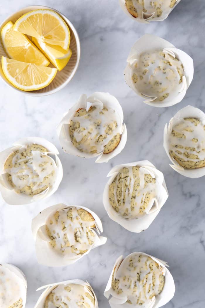 Glazed lemon poppy seed muffins in their wrappers scattered on a marble countertop.