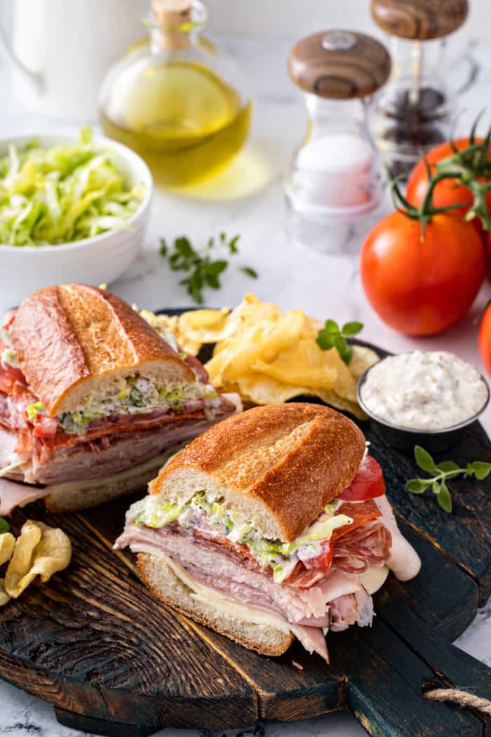 Halved grinder sandwich set on a wooden serving board next to potato chips and a ramekin of the sandwich dressing.