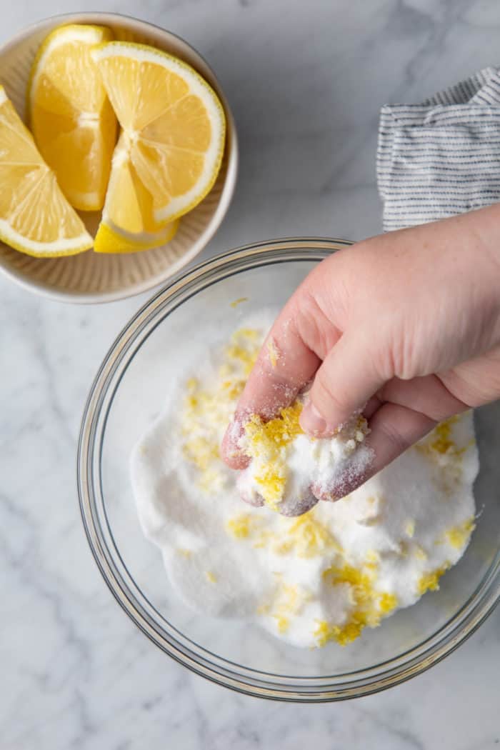 Hand rubbing together granulated sugar and lemon zest in a glass bowl.