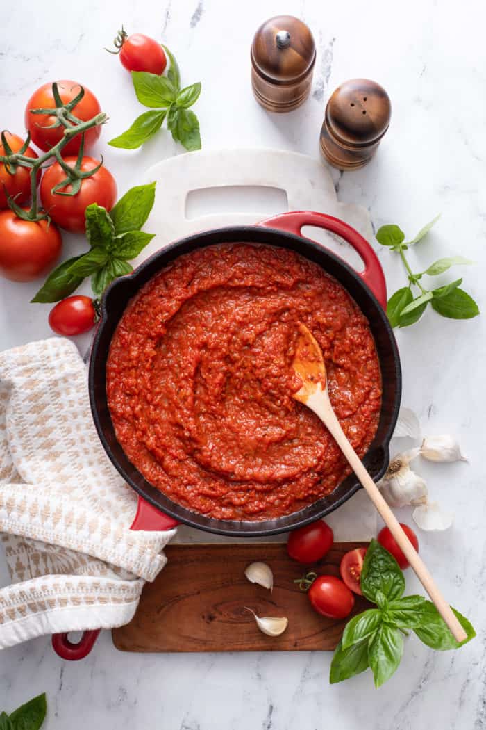 Quick tomato sauce in a skillet, set on a wood and marble board and surrounded by tomatoes, garlic, and basil.