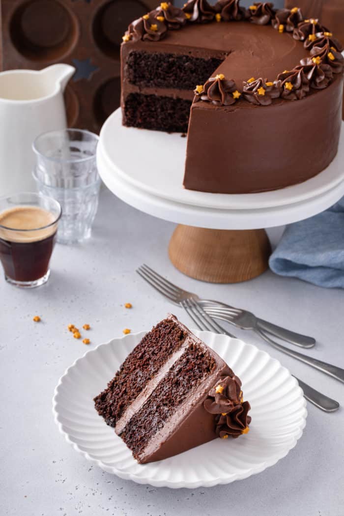 Slice of black magic cake on a white plate. A cake stand with the rest of the cake is in the background.