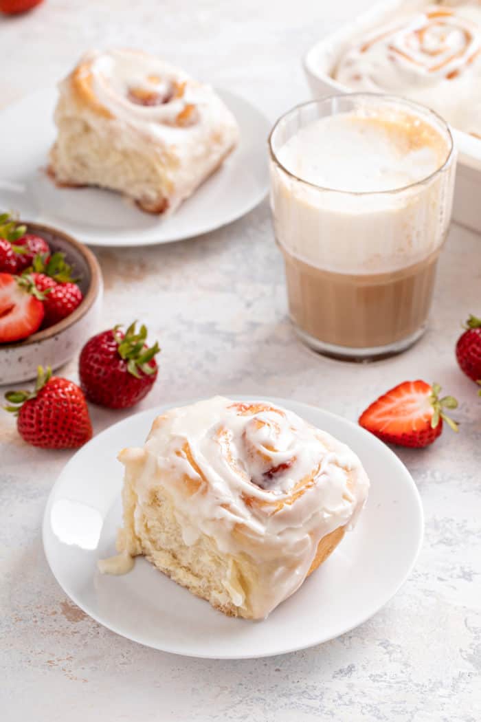 Strawberry roll topped with cream cheese frosting set on a white plate, with coffee and fresh strawberries in the background. 