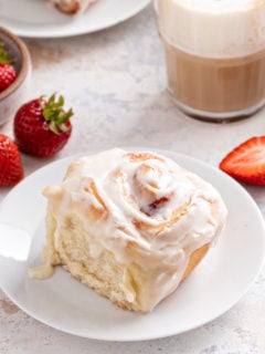 Strawberry roll with cream cheese frosting set on a white plate. Coffee and fresh strawberries are in the background.